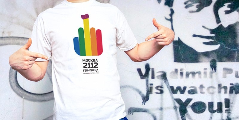 Fotografía en plano corto de un chico con una camiseta con el logotipo del orgullo gay de Moscú 2112 delante de un grafiti que dice Vladimir Putin is watching you.