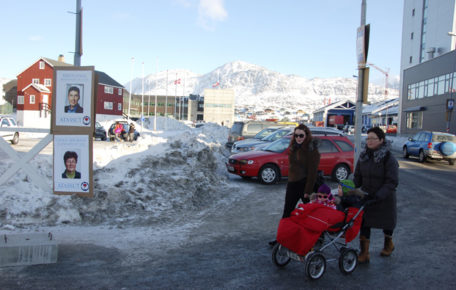 Fotografía de una calle de la capital, Nuuk, con soy y nieve, carteles electorales, dos mujeres Inuit y un bebé en un carrito.