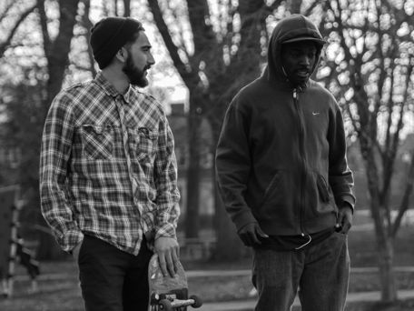 g0y bromance, black and white photo of two friends talking while walking in the park, one of them is a skateboarder.